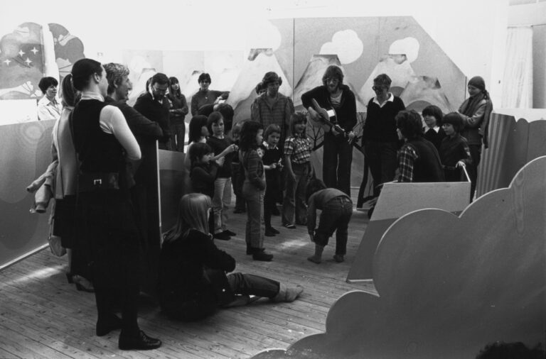 En grupp med vuxna och barn står samlade inne i konsthallen. En av de vuxna ser ut att spela gitarr.
