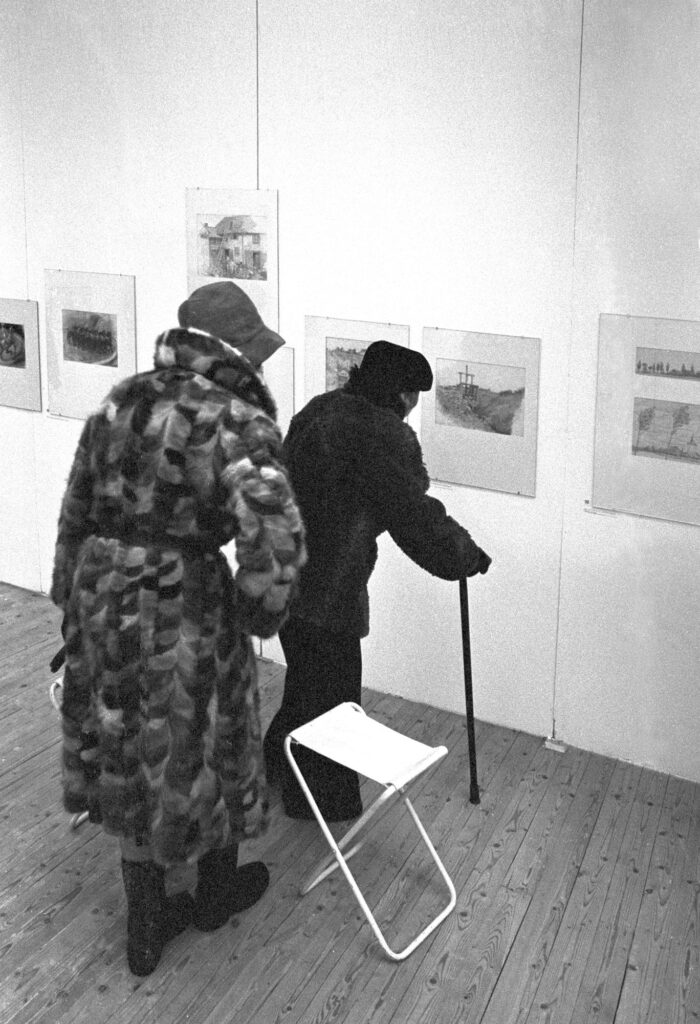 Två personer fotograferade bakifrån och lite ovanifrån. En person använder käpp. Personerna tittar på konst uppsatt på väggen. Bredvid dessa står en fällstol.