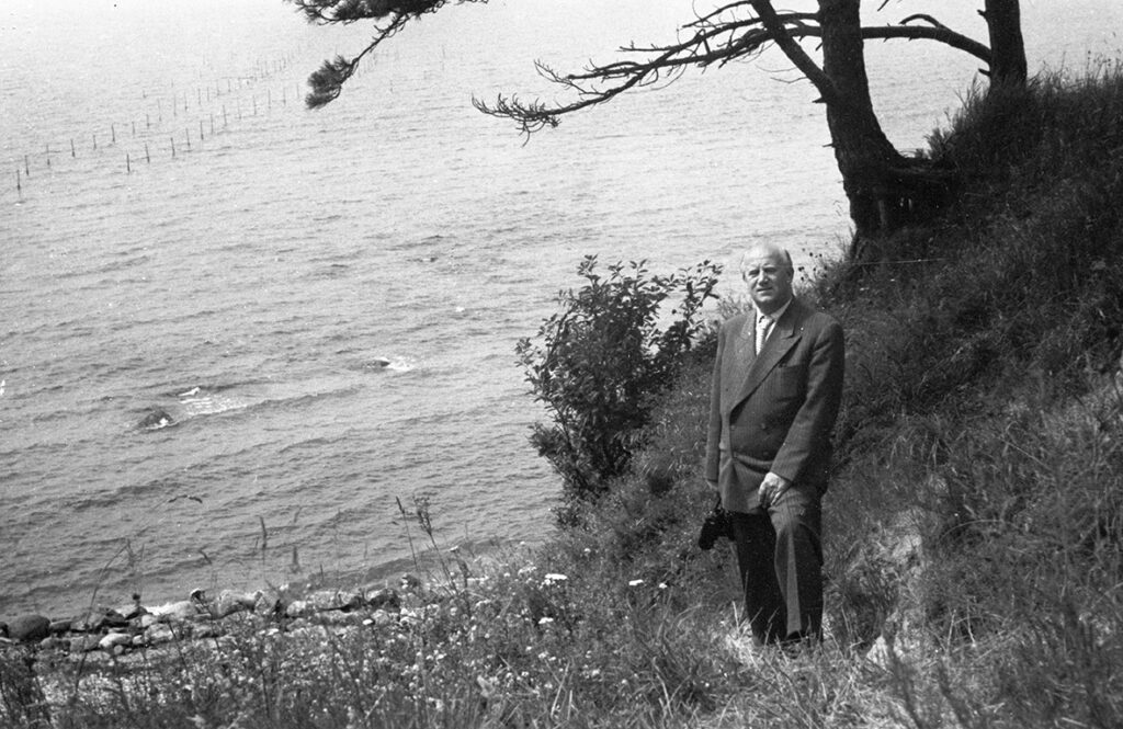 En man poserar för kameran i en blommande slänt ned mot havet.