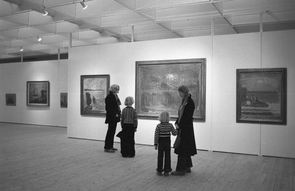 Tavlor på väggarna i utställningshallen. Två vuxna och två barn står och kollar på en av de större tavlorna.