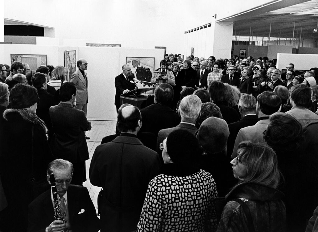 En överblicksbild från öppningen av konsthallen. Det är överfullt med många besökare. Mitt i massan av människor syns en man stå i en talarstol. Publiken är vänd mot honom.