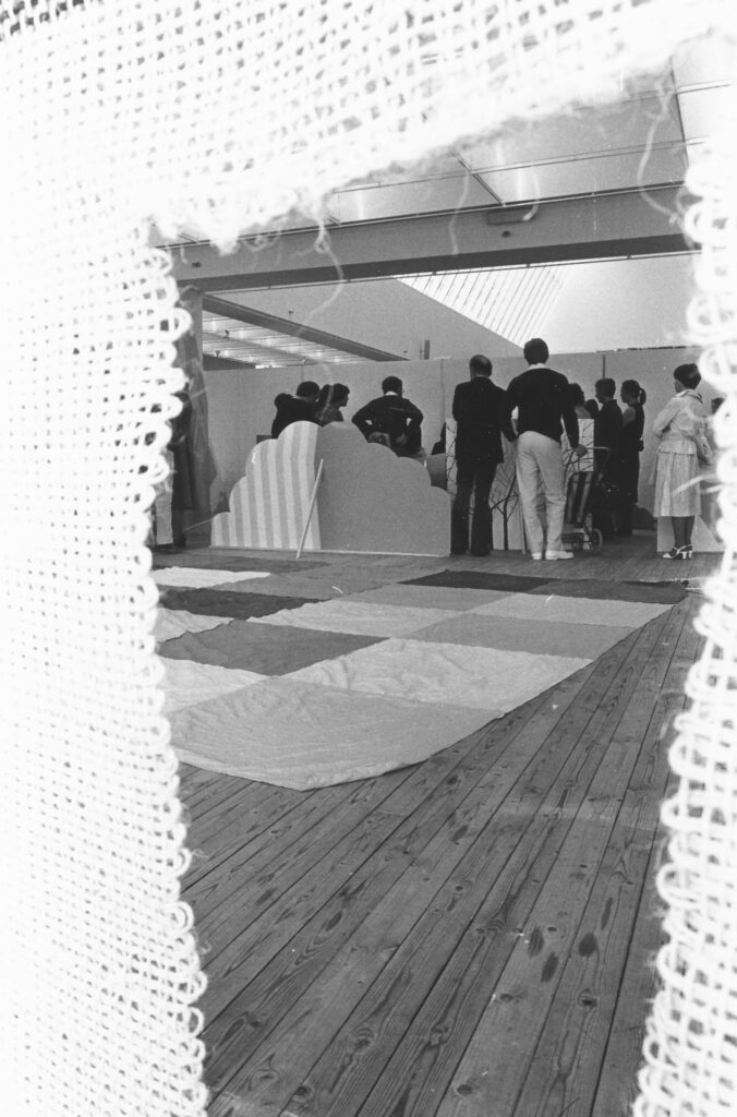En grupp personer står samlade inne i en konsthall, fotograferade genom ett virkat ljust tyg. På golvet framför personerna ligger en stor filt i rutigt mönster.