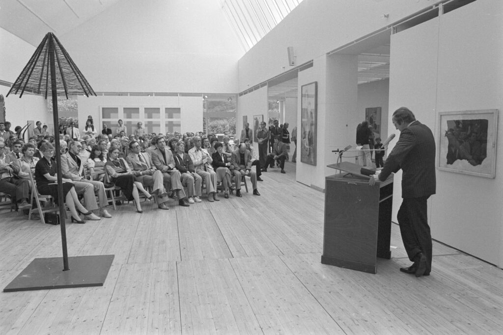 Ett svartvitt foto av en stor publik som sitter i en konsthall och lyssnar till en man som står och talar vid ett podium. Hallen har ljust trägolv och vita väggar, där flera konstverk är placerade.