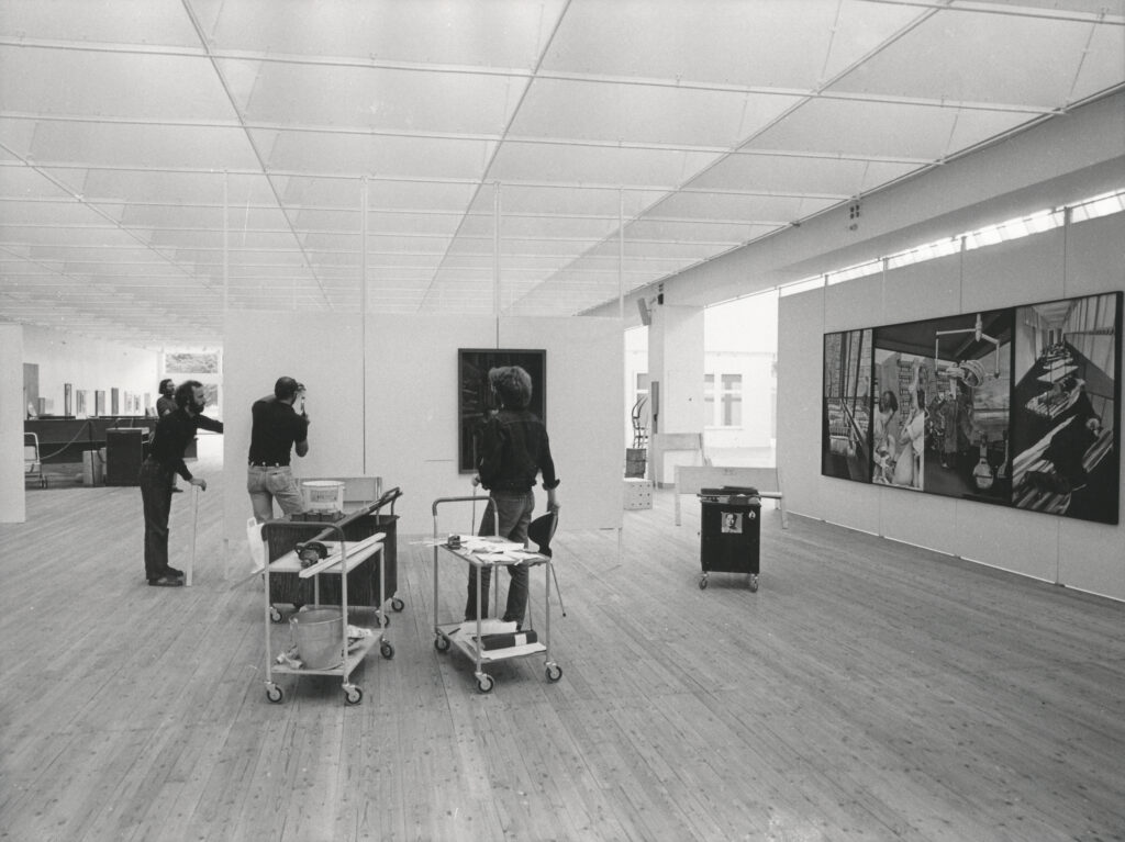 Tre personer installerar konstverk i en konsthall. En står framför en vägg och håller i en hammare. Konsthallen har ljusa väggar och trägolv.