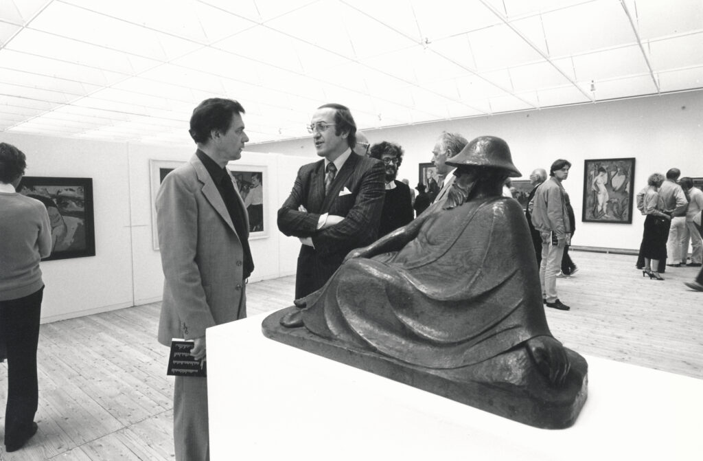 En grupp män står och samtalar i en konsthall. I bakgrunden rör sig besökare som