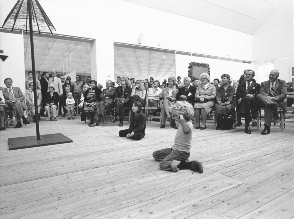 En stor publik sitter i en konsthall och lyssnar till en man som står och talar vid ett podium. Hallen har ljust trägolv och vita väggar, där flera konstverk är placerade.