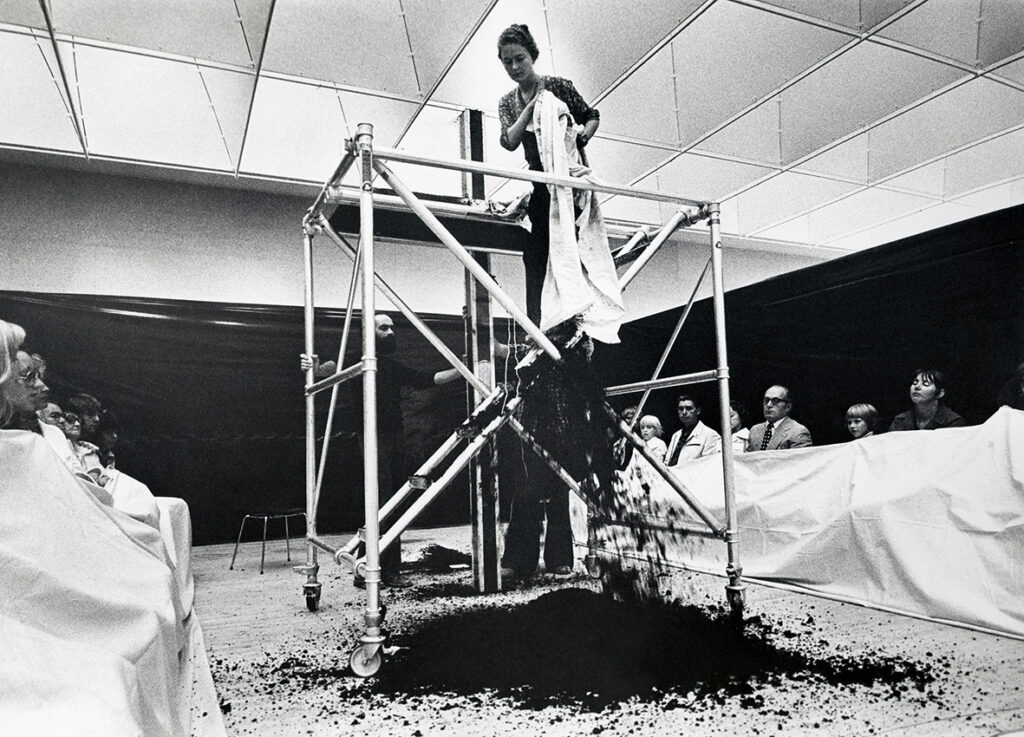 Black and white image of a woman in a performance. She stands on a platform, spreading soil on the floor.