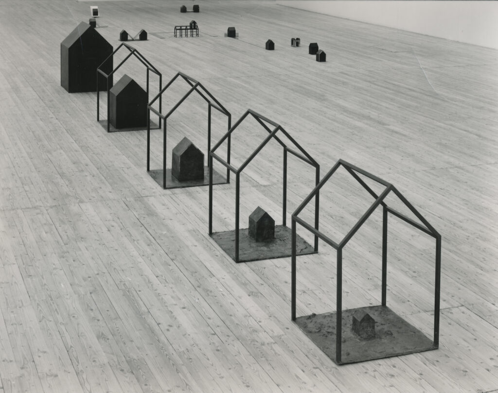Skulpturer formade som hus, utplacerade på ett ljust trägolv i en konsthall.