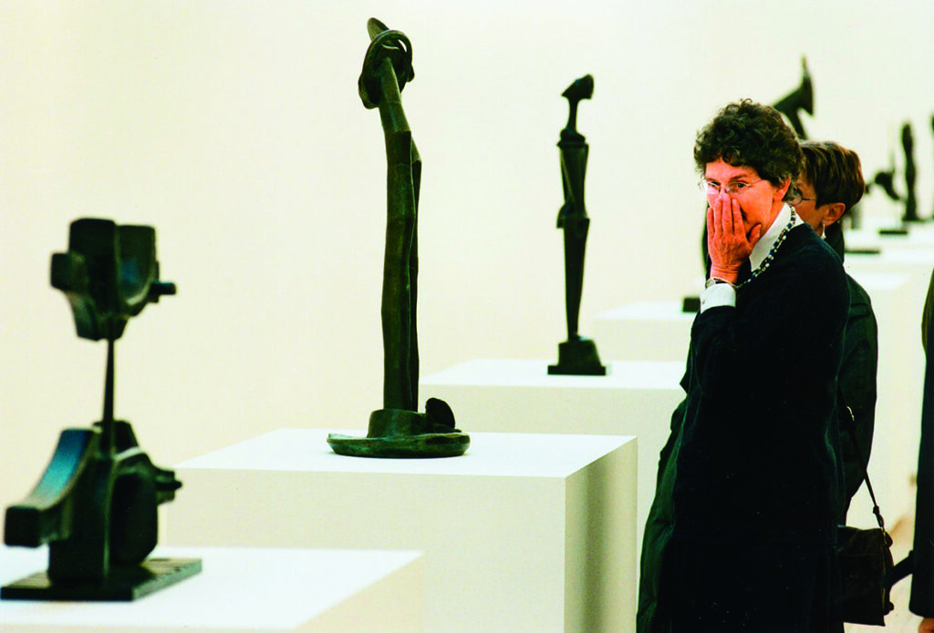 Flera skulpturer står uppradade på piedestaler. Till höger i bild står en kvinna med kortklippt mörkt hår och glasögon, hon lutar hakan i handen och kollar på verken.