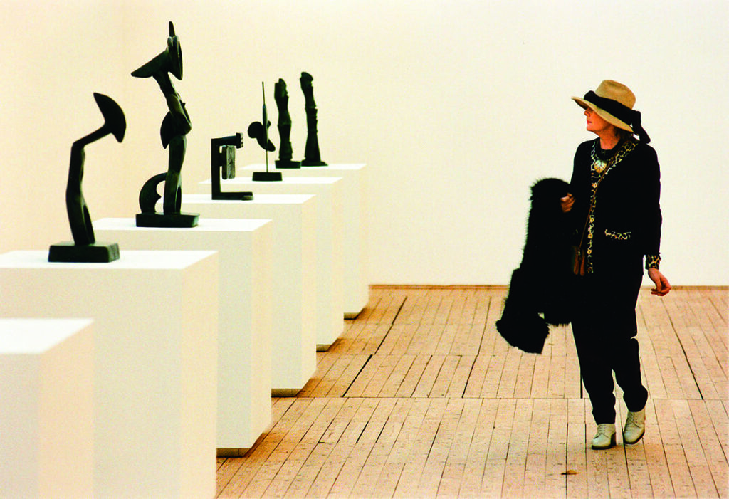 Flera skulpturer står uppradade på piedestaler. Till höger i bild går en kvinna med stor solhatt och bär sin kappa över armen. Hon kollar på skulpturerna.