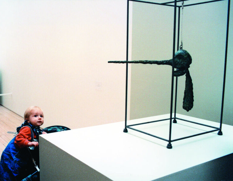 Skulptur av Alberto Giacometti hängandes i en ställning, till vänster i bild syns ett litet barn som kollar upp på skulpturen från sin barnvagn.