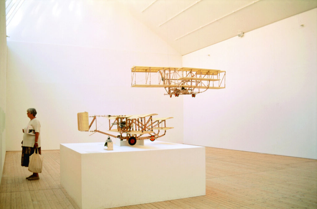 Anders Österlins verk, två stycken träkonstruktioner som liknar stora flygplan. Ett av dem hänger i taket och det andra står på en stor piedestal. Till vänster i bild syns en person som betraktar någonting som hänger på väggen intill.