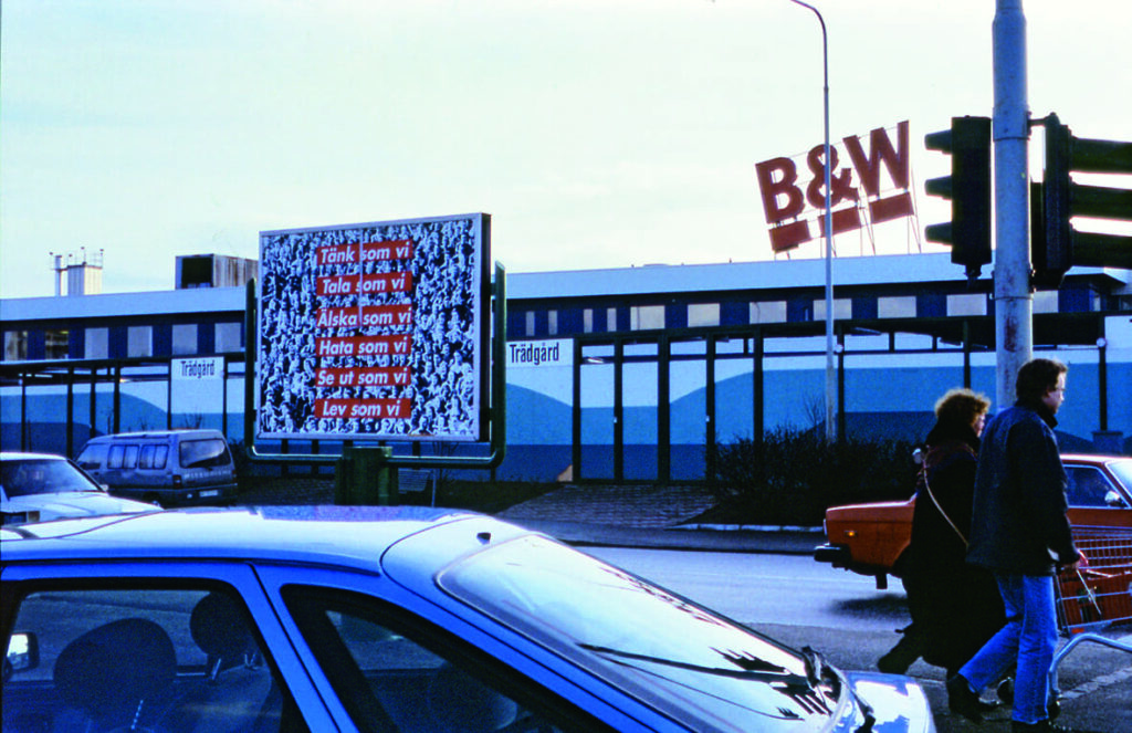 På en skylt vid en bilväg finns Barbara Kreugers kollage. I förgrunden står en bil parkerad och till höger i bild går ett par med en kundvagn över ett övergångsställ.