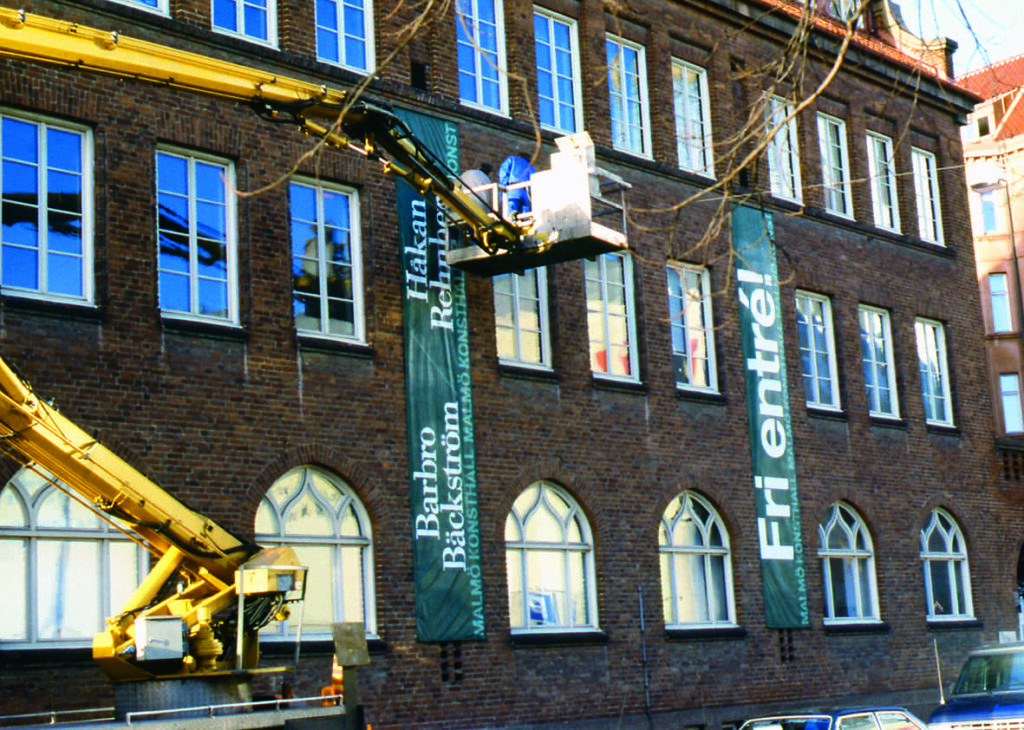 Två personer står i en skylift och installerar två långa tyger som hänger ner för museets fasad för att marknadsföra Barbro Bäckströms utställning. På ena står det Barbro Bäckström, Malmö Konsthall. Och på den andra står det fri entré!