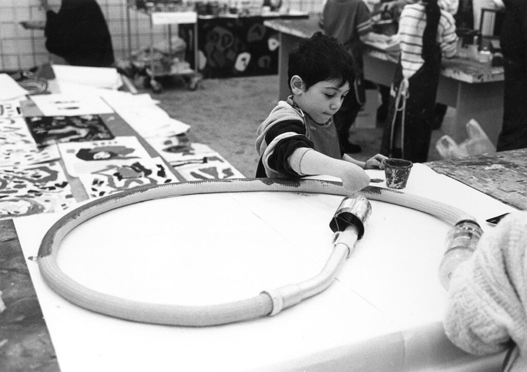 En pojke målar en slang med pensel, han har mörkt hår och kollar koncentrerat på slangen. I bakgrunden syns andra barn ståendes runt ett bord samt teckningar liggandes på ett bord intill.