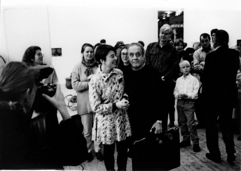 Bertil Lundberg står omringad av ett tiotal personer inne i konsthallen. Han håller en väska i ena handen och bredvid honom står en kvinna med kort mörkt hår och ljus, mönstrad klänning.