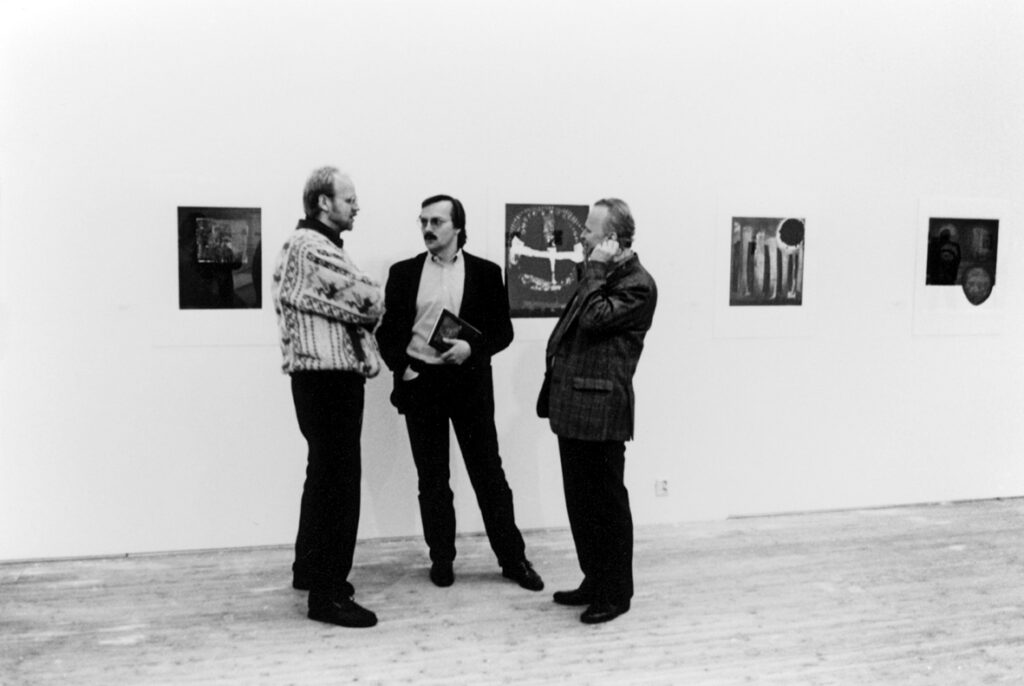 Tre män står och pratar med varandra framför en vägg med tavlor.