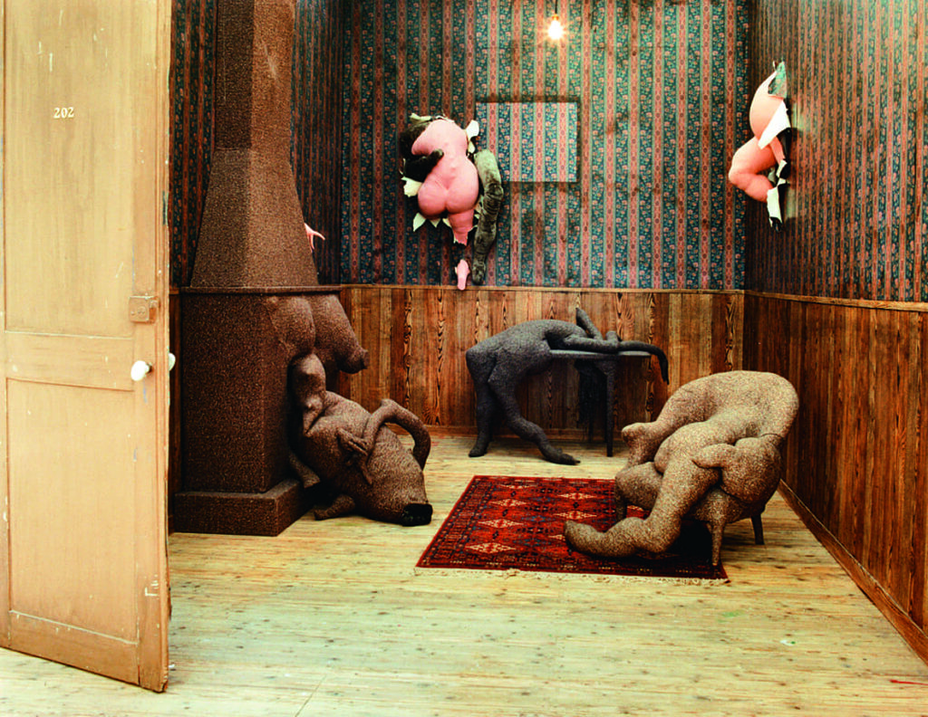 Ett av Dorothea Tannings verk. Ett rum med randig tapet där abstrakta former i rosa material böljer ut från väggarna. Två människoliknande former i brun färg står på golvet och en slank form i svart ligger över ett litet bord.
