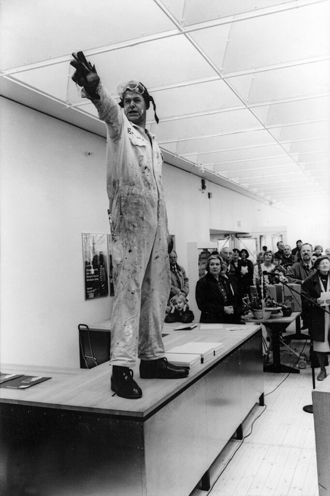 En person står på en bänk i Konsthallen iklädd en smutsig, ljus overall och pekar ut i luften. Runtom står personer och tittar på.