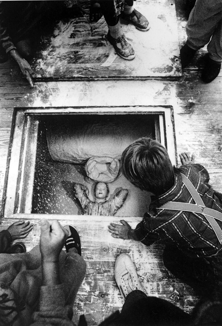 En liten pojke med hängselbyxor tittar ner genom en lucka i golvet, där nere ligger en person som blundar.