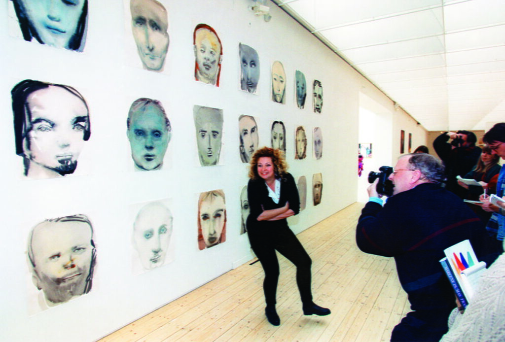Marlene Dumas framför en vägg i konsthallen, hon poserar med armarna i kors och ler stort. Till höger i bild syns flera fotografer som tar bilder av Marlene.