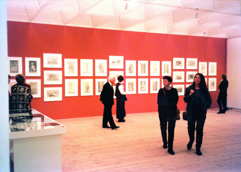 Flera personer går runt inne i konsthallen, i bakgrunden syns en röd vägg med flera målningar upphängda.