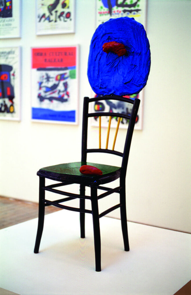 Ett av Joan Mirós verk. En svart stol med grönmålad sits och gula pinnar i ryggstödet. Ovanpå ryggstödet står en skulptur av en blå rund form med ett rött organiskt utstick.