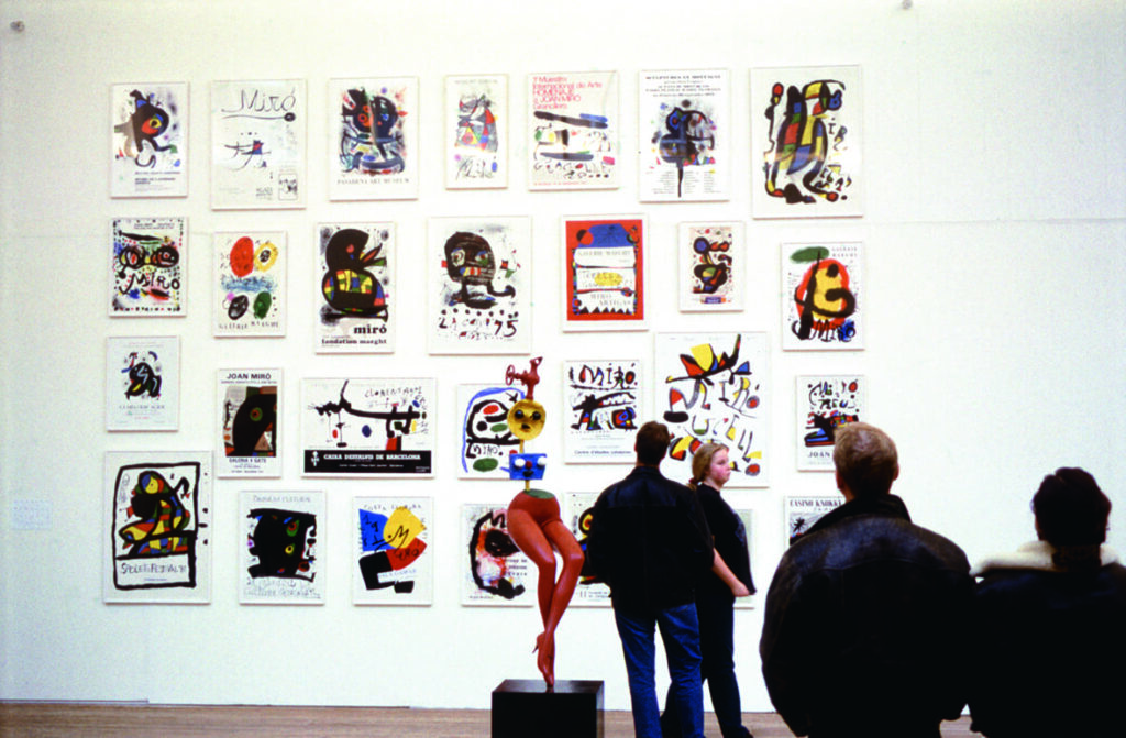 Miljöbild över Joan Mirós affischer. Cirka 30 stycken affischer hänger inramade på en vit vägg. I förgrunden syns fyra personer stå och betrakta verken.