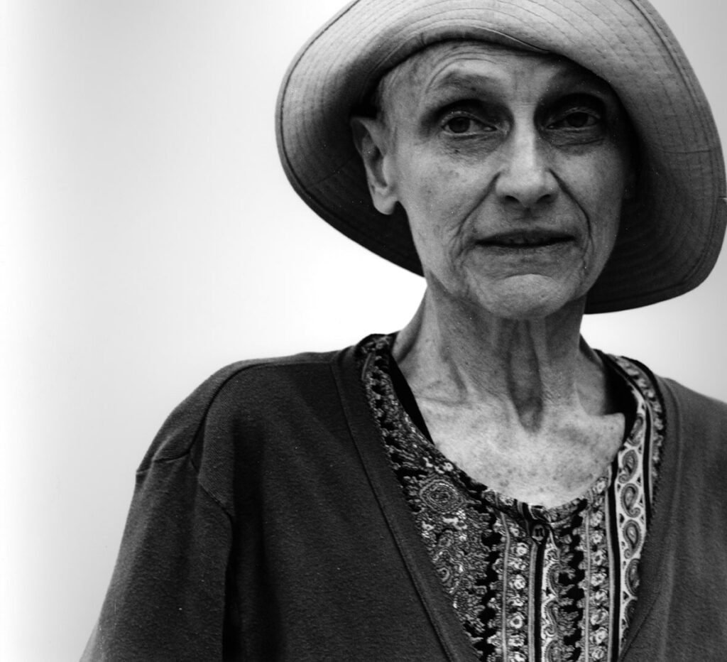 Svartvit bild av Nancy Spero, hon är iklädd en mönstrad tröja med en mörk kofta över. På huvudet bär hon en hatt i mjukt material med ena kanten uppvikt.