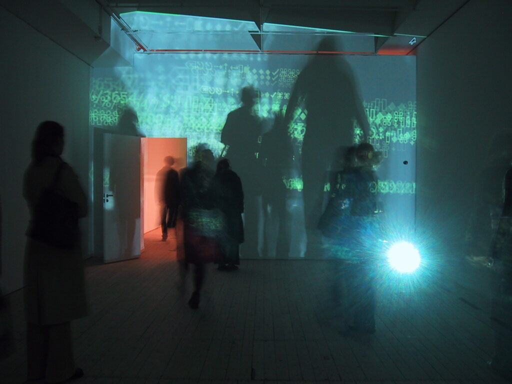 Installationsbild från utställningen Electrohype 2002. Ett dunkelt rum med flera besökare och en projektion med siffror och symboler i grönt.