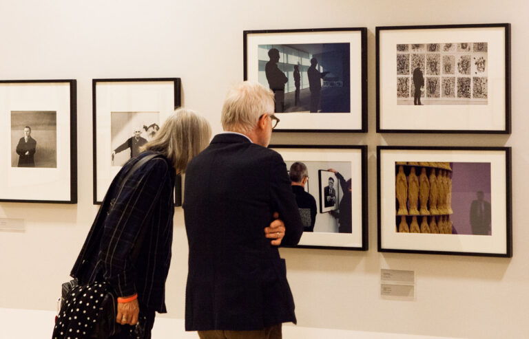 Stämningsbild av utställningen Malmö Konsthall 40 år, 2015. Några besökare tittar närmre på fotografier som sitter inramade på väggen.