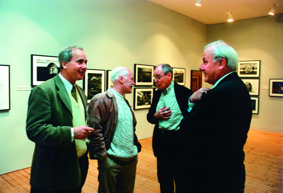 Four men are standing and talking in front of a wall full of paintings. They are all wearing blazers.