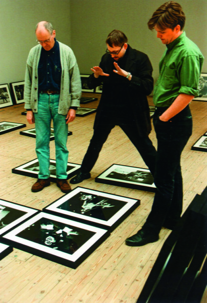 Tre män står och diskuterar tavlor de lagt ut på golvet inför hängning. Killen längst till höger är iklädd grön skjorta, mannen i mitten har helsvart på sig och han till vänster har mörkblå skjorta, grå kofta och jeans.