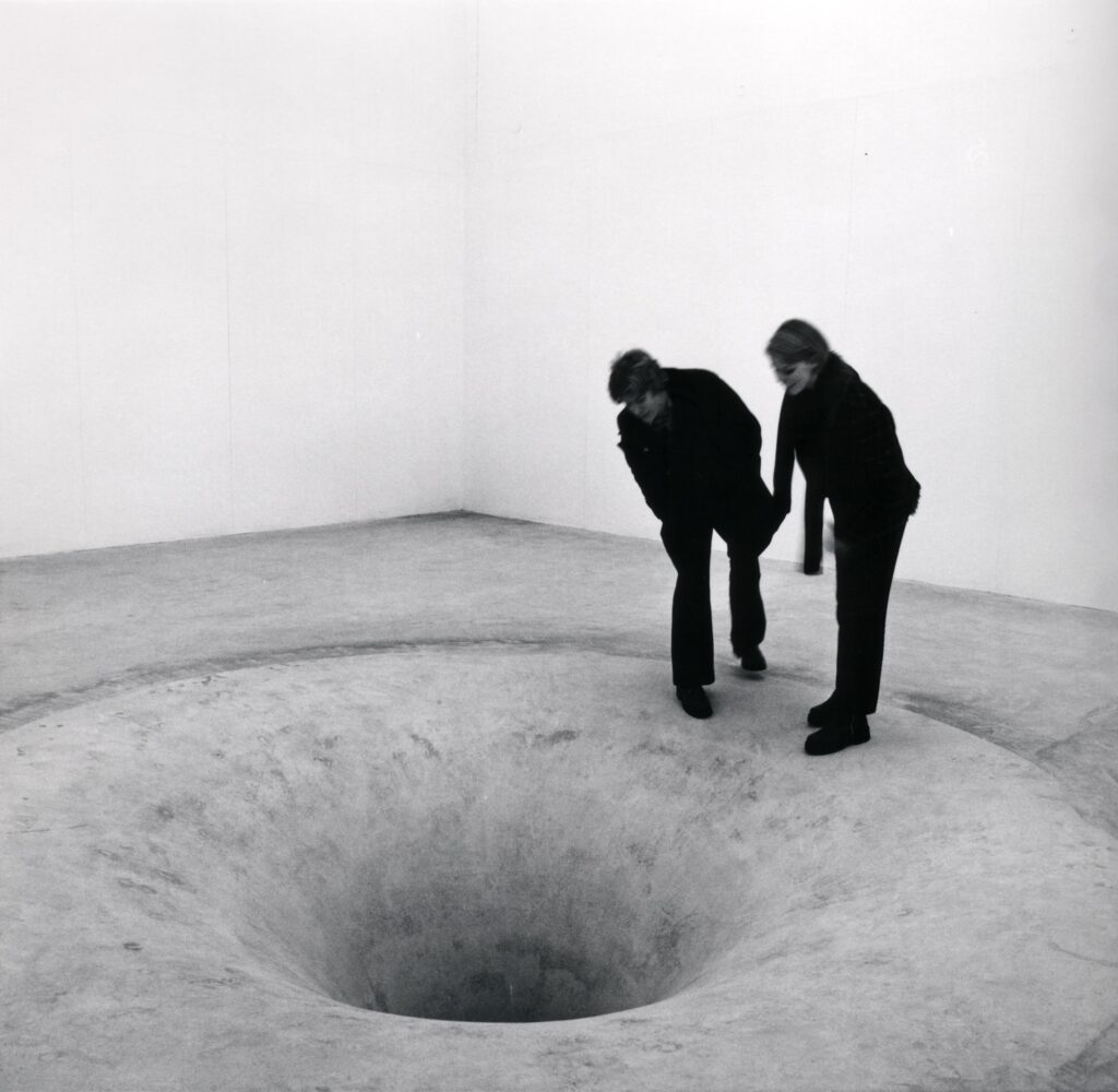 Svartvit bild av personer som kikar ned i ett till synes bottenlöst betonghål i konsthallens golv.