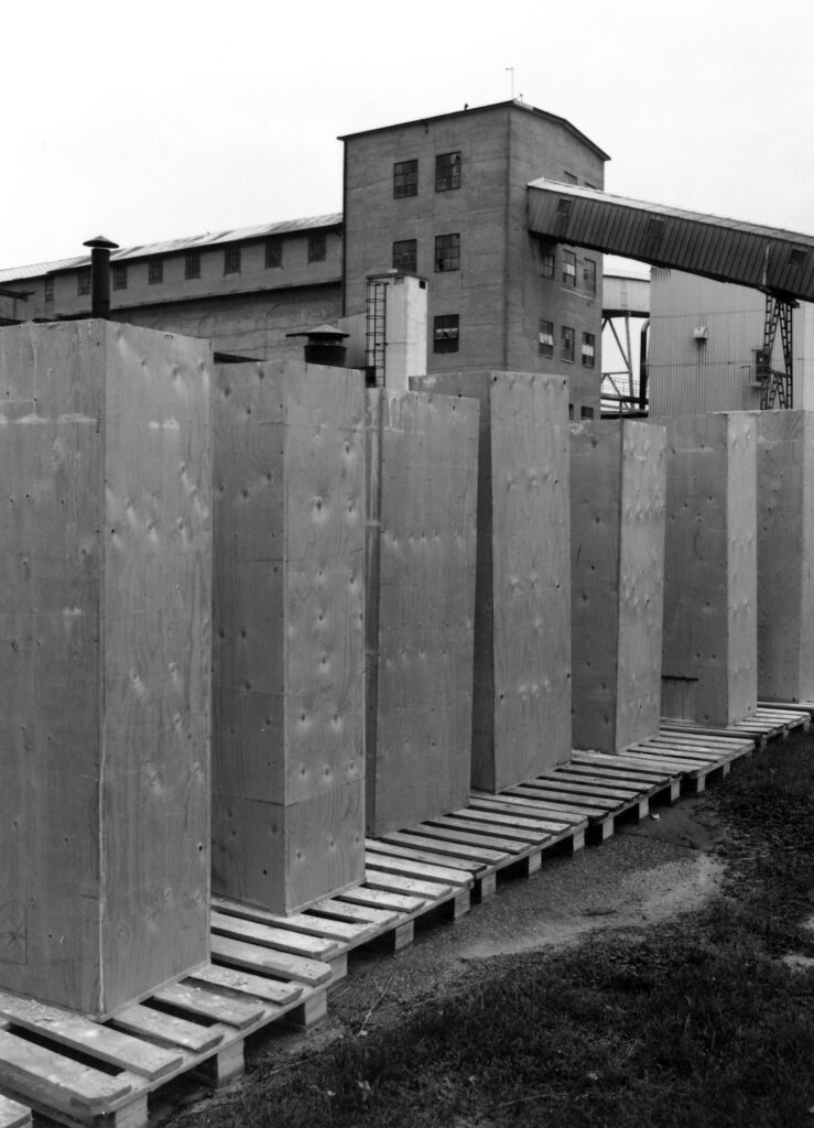 Industrimiljö med en rad stora, stående pelarliknande trälådor placerade på lastpallar i förgrunden. I bakgrunden syns en fabrik med flera fönster och skorstenar.