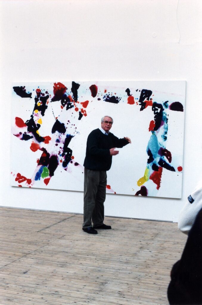En man står med ryggen mot en färgstark målning i en konsthall. Mannen gör en gest med handen.