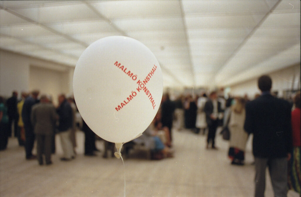 Stämningsbild från konsthallen med massor av suddiga besökare i bakgrunden och en ballong med Malmö Konsthalls logga i förgrunden.