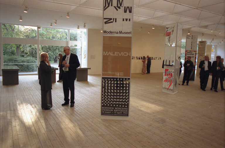 Installationvy av utställningen "John Melin – till exempel". Bilden är tagen under vernissage eller någon tillställning med besökare spridda i eftermiddagsljuset.