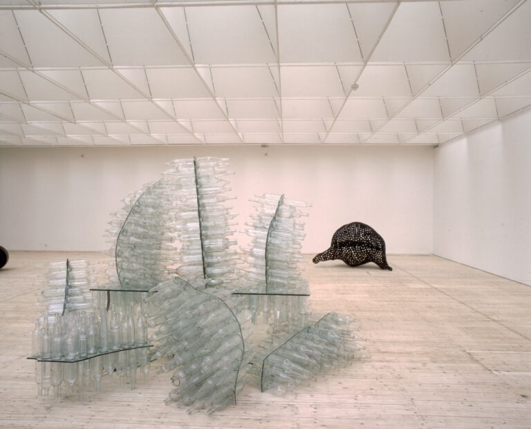 En installation inne i en konsthall, bestående av en skulptur uppbyggd av staplade glasflaskor i en böljande, organisk form. I bakgrunden syns en svart, prickig skulptur som liknar en djurlik form.