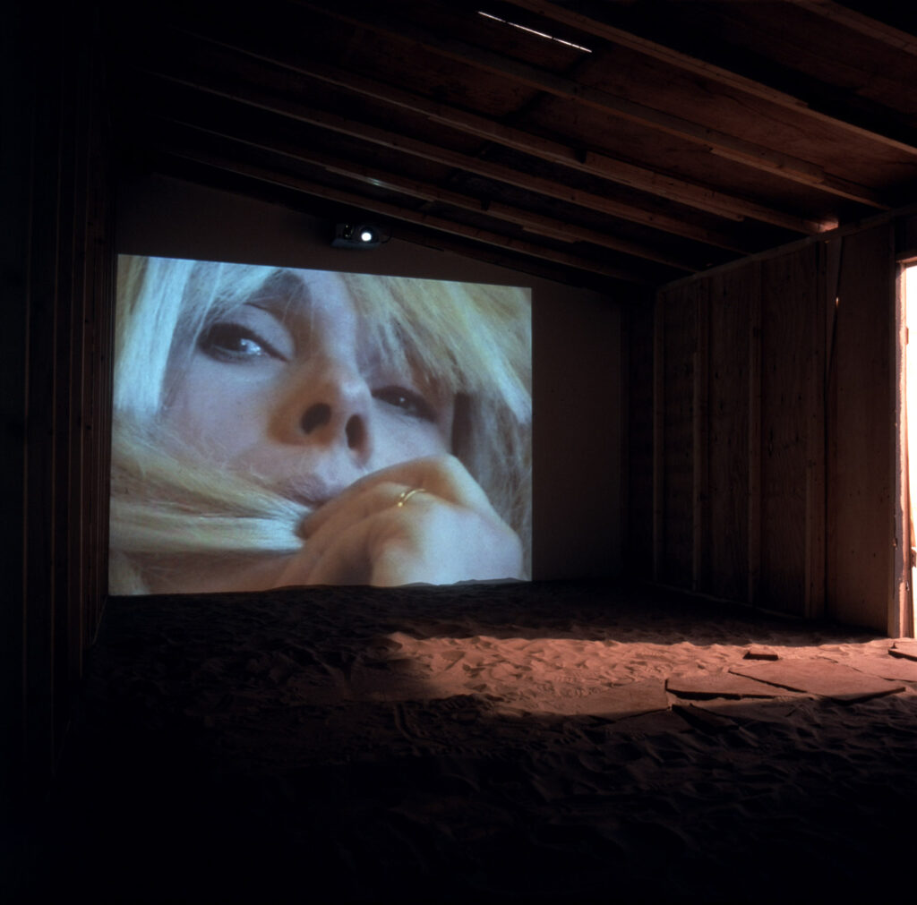 Installation av Lena Mattson. En videoprojektion, närbild, av en blond kvinnas ansikte i ett rum som påminner om ett skjul. På marken finns sand och plattor.