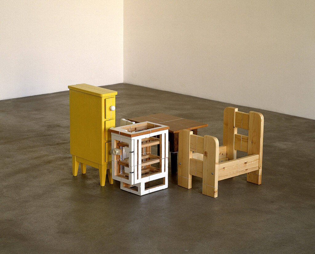 Konstverk av Axel Lieber. Ett mindre antal föremål/små möbler i trä står på golvet i ett rum. Möblerna ser krympta och dekonstruerade ut - som att de en gång varit större, men att delar har sågats bort och de sedan satts ihop igen till mindre versioner.