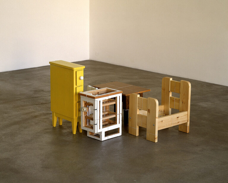 Konstverk av Axel Lieber. Ett mindre antal föremål/små möbler i trä står på golvet i ett rum. Möblerna ser krympta och dekonstruerade ut - som att de en gång varit större, men att delar har sågats bort och de sedan satts ihop igen till mindre versioner.