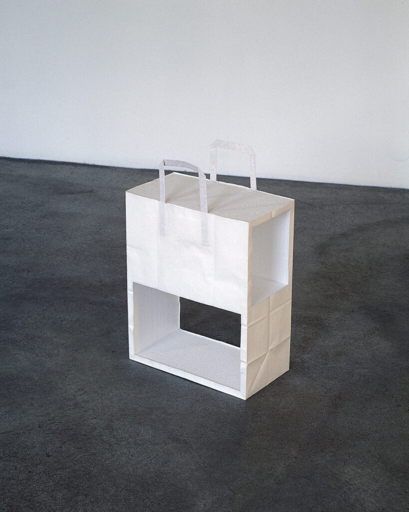 Konstverk av Axel Lieber. En vit papperskasse med handtag har skurits upp på ett arkitektoniskt vis, som att den har stora, rektangulära fönster och två våningar.