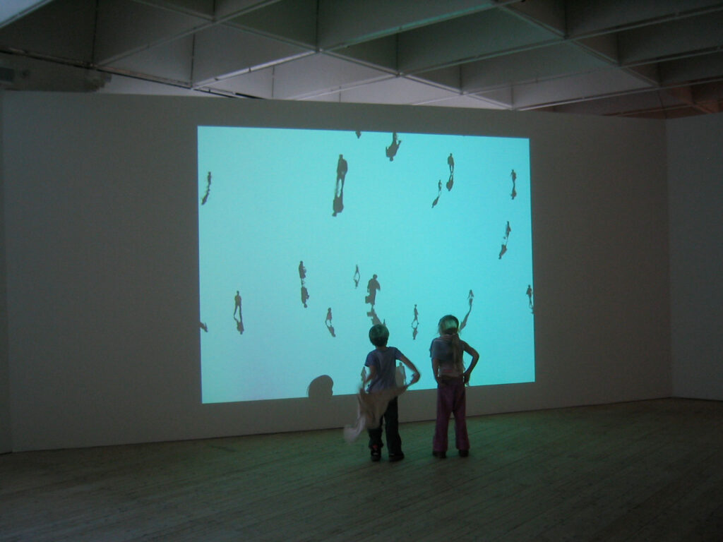 Installationsbild/vy av utställningen Electrohype 2004. Två unga besökare tittar på en projektion av promenerande små figurer.