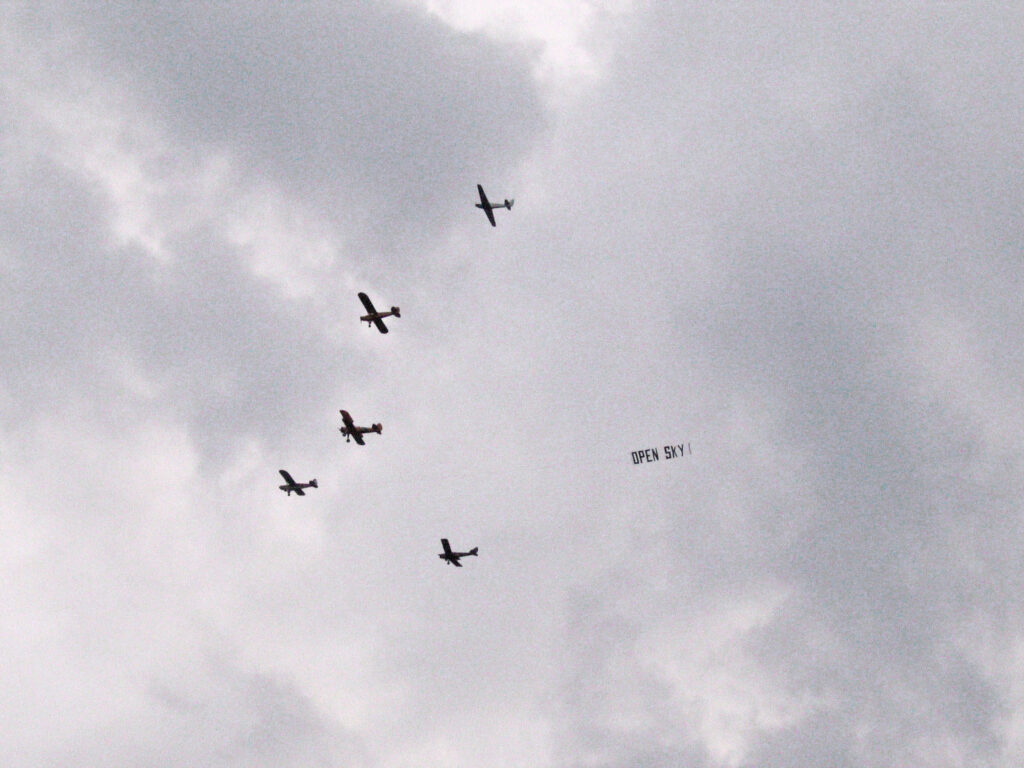 Fem stycken mindre flygplan på en molnig himmel. En bandaroll är fäst vid ett av planen med texten OPEN SKY.