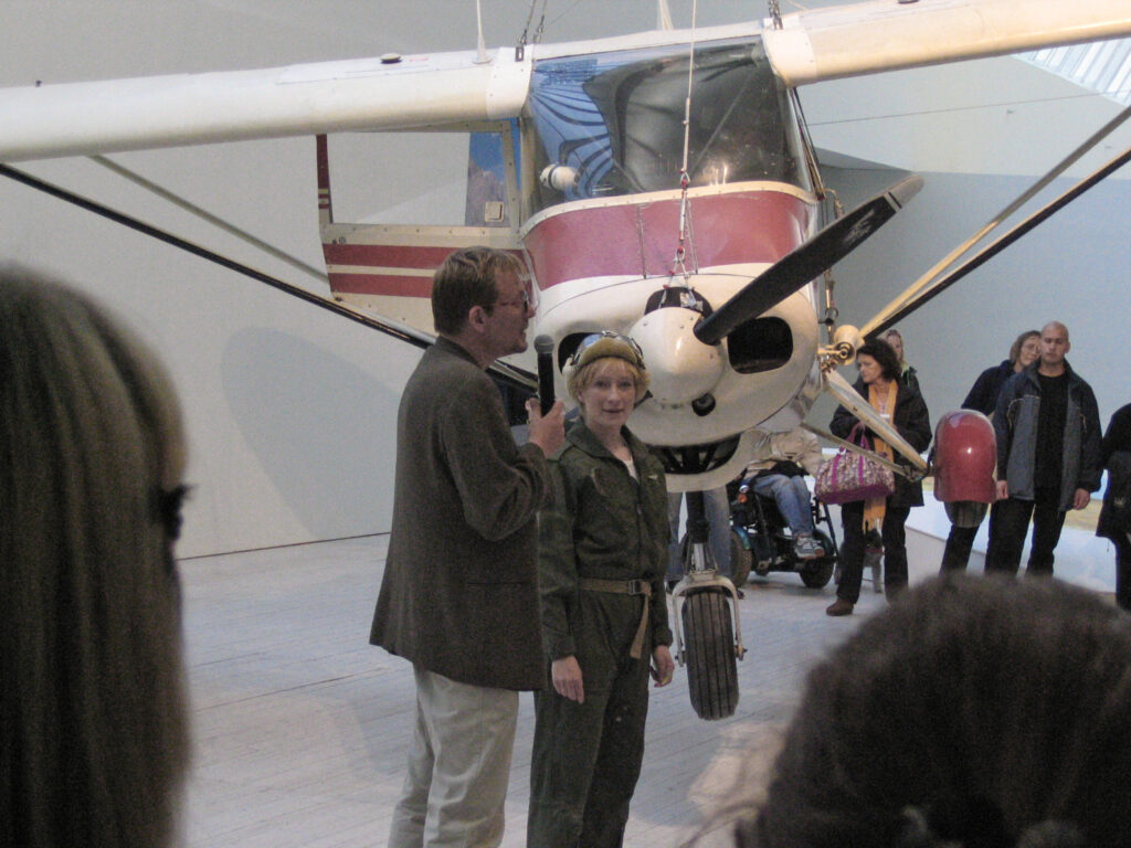 Konsntären står med flygplansmössa framför sitt flyplan som hänger från taket på Malmö Konsthall. Bredvid henne står Lars Gramby, konsthallschef och inviger utställningen.