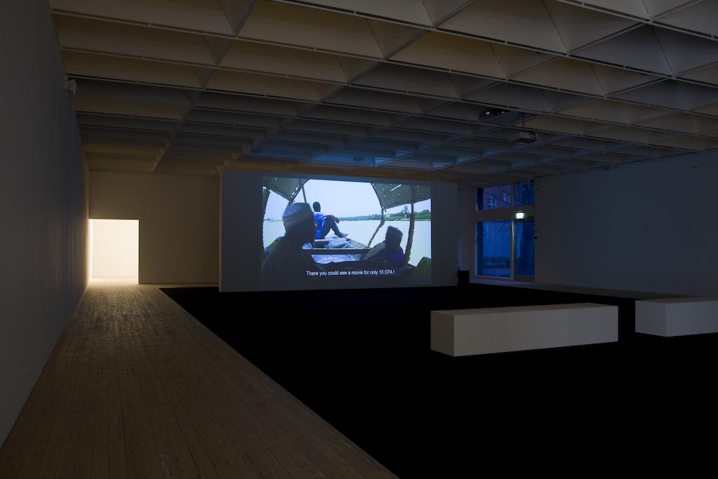 Ett rum med en vit bänk med en videoprojektion. På filmen ser man tre personer som sitter på en båt.