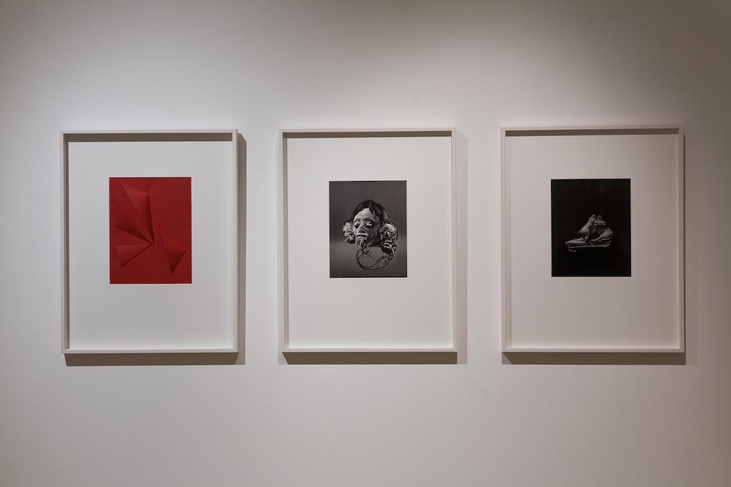Tre konstverk hänger på rad på en vit vägg. Den vänstra bilden är helt i rött, de andra två är svartvita fotografier, ett med en afrikanskliknande mask och den andra en skulptur.