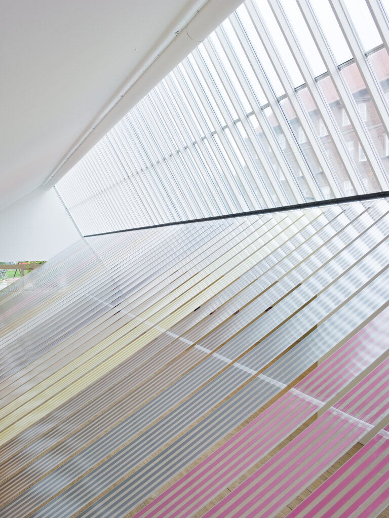 Installationsbild av breda plastremsor fyllda med kryddor som sträcker sig från ljusschaktets fönster och ned till golvet.