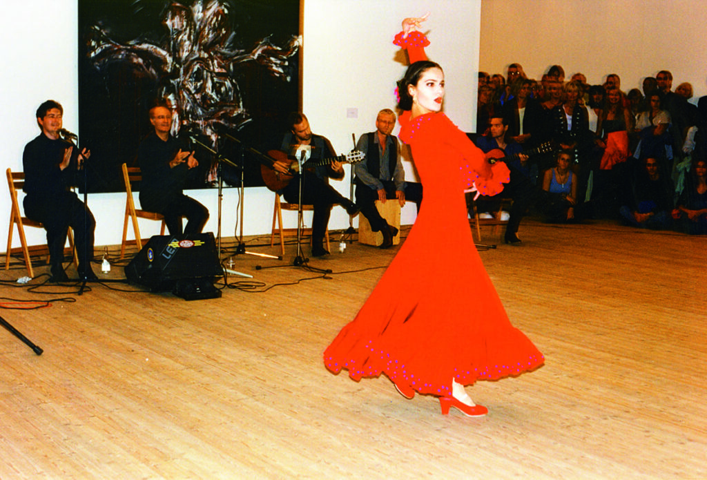 En kvinna iklädd röd klänning med stor kjol. Hon dansar och har ena armen över huvudet och den andra armen runt sin egna överkropp. I bakgrund sitter ett band, 5 män, som spelar musik.